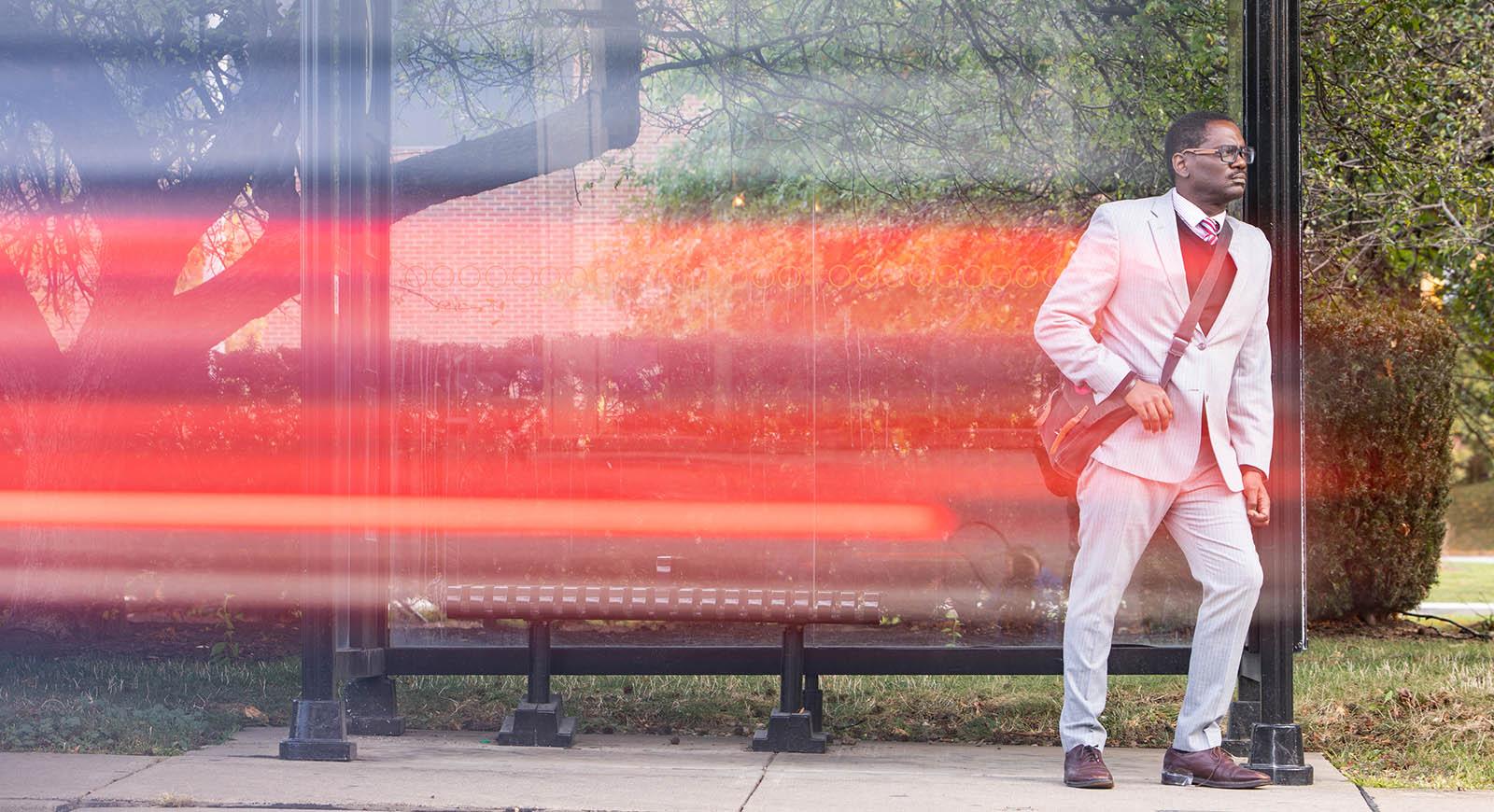 Photo of Brian Broome, a Black man in a grey suit with a messenger bag, waiting at a bus stop while a bus passes