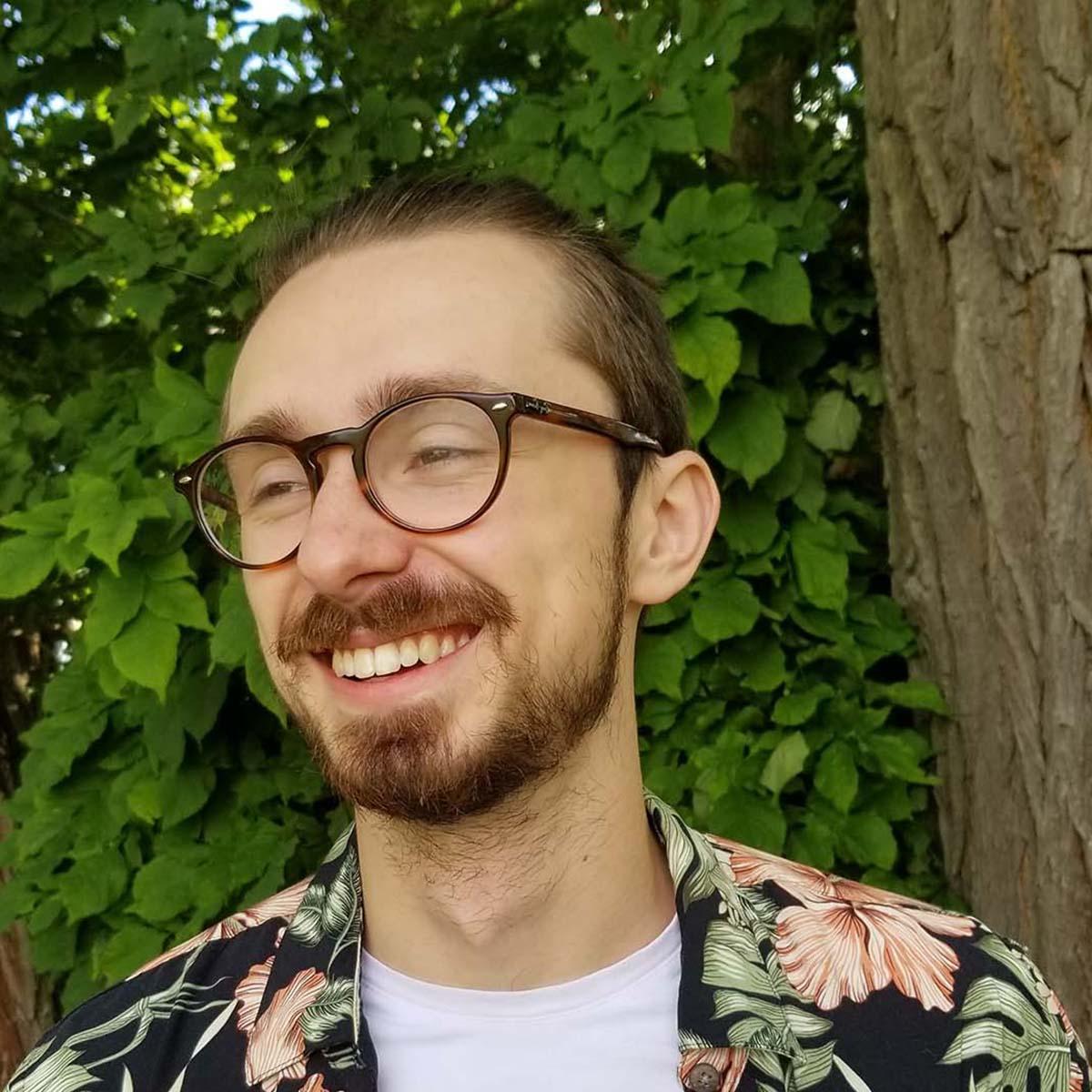 卡森·卡斯特的照片, a young white man with glasses and a bright floral shirt, 在绿树前微笑